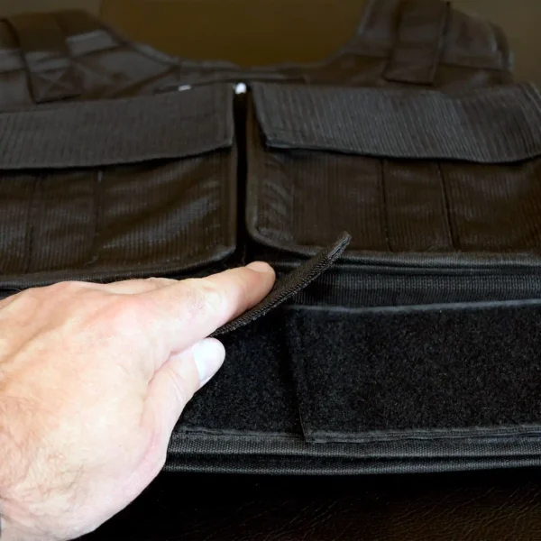 An individual adjusts the Velcro strap on an Ironmaster Ultimate Training Vest lying on a dark surface. This black weighted vest features multiple pockets and zippers, with the person's hand visible on the left side of the image.