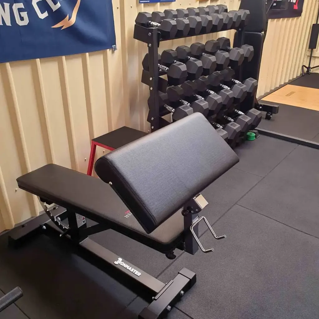 A gym interior featuring an Ironmaster Super Bench PRO with an arm pad and a rack filled with various dumbbells. The floor is covered with black mats, and part of a wall with a banner is visible in the background, enhancing the workout atmosphere.