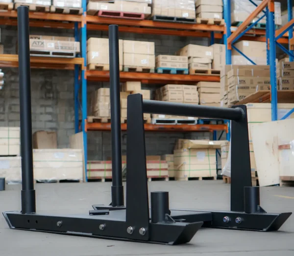A Power Maxx Gym Weight Sled with vertical handles rests on the warehouse floor, where shelves brimming with cardboard boxes are visible in the background.