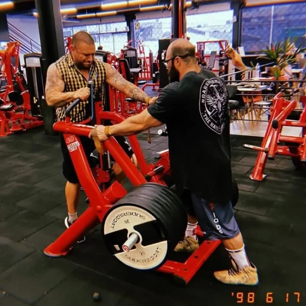 Two men at a gym; one wears a plaid vest and guides the other in a black shirt and shorts using a weight-lifting machine with multiple plates. The gym, outfitted with various machines and exercise gear, includes the GymPin 2 Internal Extender (Not Pair).