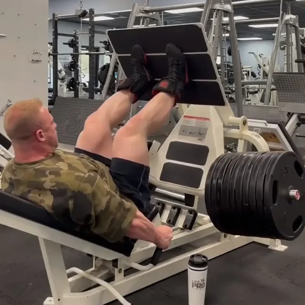 A person in the gym uses the GymPin 2 Internal Extender for leg presses with heavy weights. They wear a camouflage shirt and black shorts, and a protein shaker is on the ground nearby, with gym equipment visible in the background.