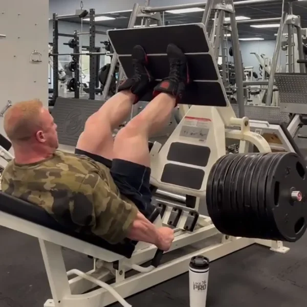 A person in the gym uses the GymPin 2 Internal Extender for leg presses with heavy weights. They wear a camouflage shirt and black shorts, and a protein shaker is on the ground nearby, with gym equipment visible in the background.