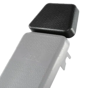 Close-up of two gym bench pads with textured surfaces, one black and square, slightly elevated above the gray one. The gray pad, labeled ATX, is part of the ATX® Headrest Extension PU.