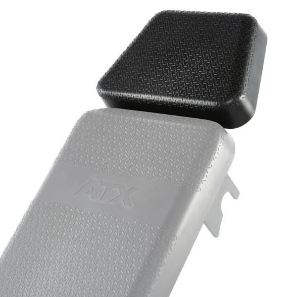 Close-up of two gym bench pads with textured surfaces, one black and square, slightly elevated above the gray one. The gray pad, labeled ATX, is part of the ATX® Headrest Extension PU.