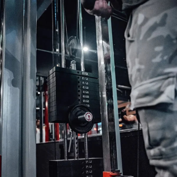 A person in camouflage pants is using a GymPin 2 Original with a cable machine at the gym. The focus is on the weight stack and handle amidst a dimly lit, intense workout setting.