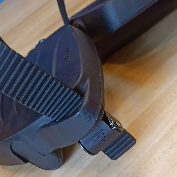 Close-up of the Spin Bike Pedal MATRIX 9/16 with a textured black strap on a light wood surface, featuring an adjustment knob for securing the foot and enhancing stability during use.
