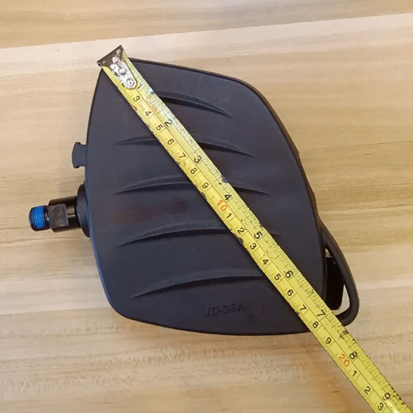The Spin Bike Pedal MATRIX 9/16 (Copy) in black is positioned on a wooden surface with a yellow measuring tape laid diagonally across it to show its size.