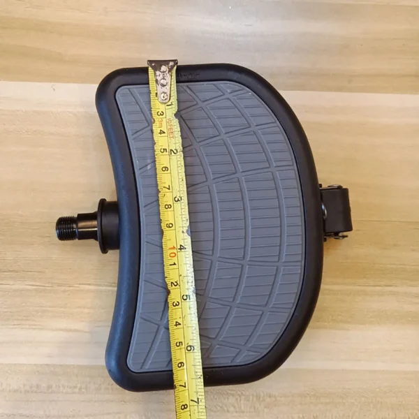 The Spin Bike Pedal 9/16 (Copy), featuring a black frame and textured gray surface, rests on a wooden surface with a yellow measuring tape across it, indicating its length is about six inches.