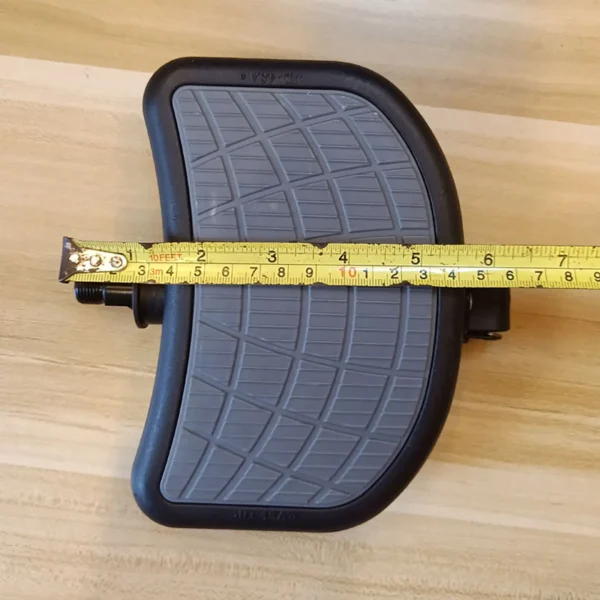 A gray Spin Bike Pedal 9/16 with a textured rubber surface and black frame rests on wood, while a yellow measuring tape indicates the pedals width is about 7 inches.