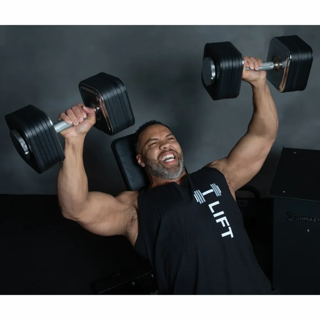 A person with a beard is performing a dumbbell bench press. They are wearing a black sleeveless shirt with the word LIFT on it. The background is dark and out of focus.