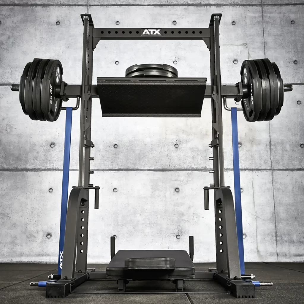 A heavy-duty squat rack with multiple weight plates on each side in a gym setting. The metal rack is set against a concrete wall background, and the floor is covered with rubber gym flooring.