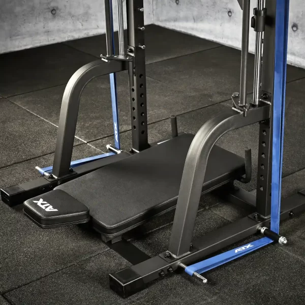 A close-up of a gym smith machine with a padded bench, metal frame, and adjustable safety bars. The floor is covered with black rubber mats, and the walls have a concrete finish. The equipment is branded with ATX logos.