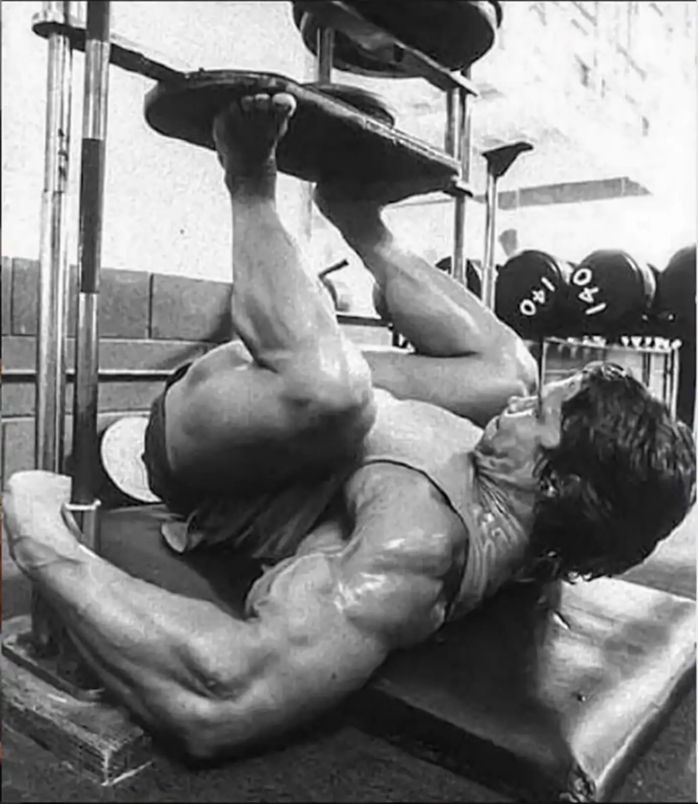 A muscular man exercises intensely on the ATX® Vertical Leg Press at the gym. Shirtless, he showcases his toned muscles and unwavering focus. In the black-and-white scene, dumbbells in the background enhance the intensity of his workout session.